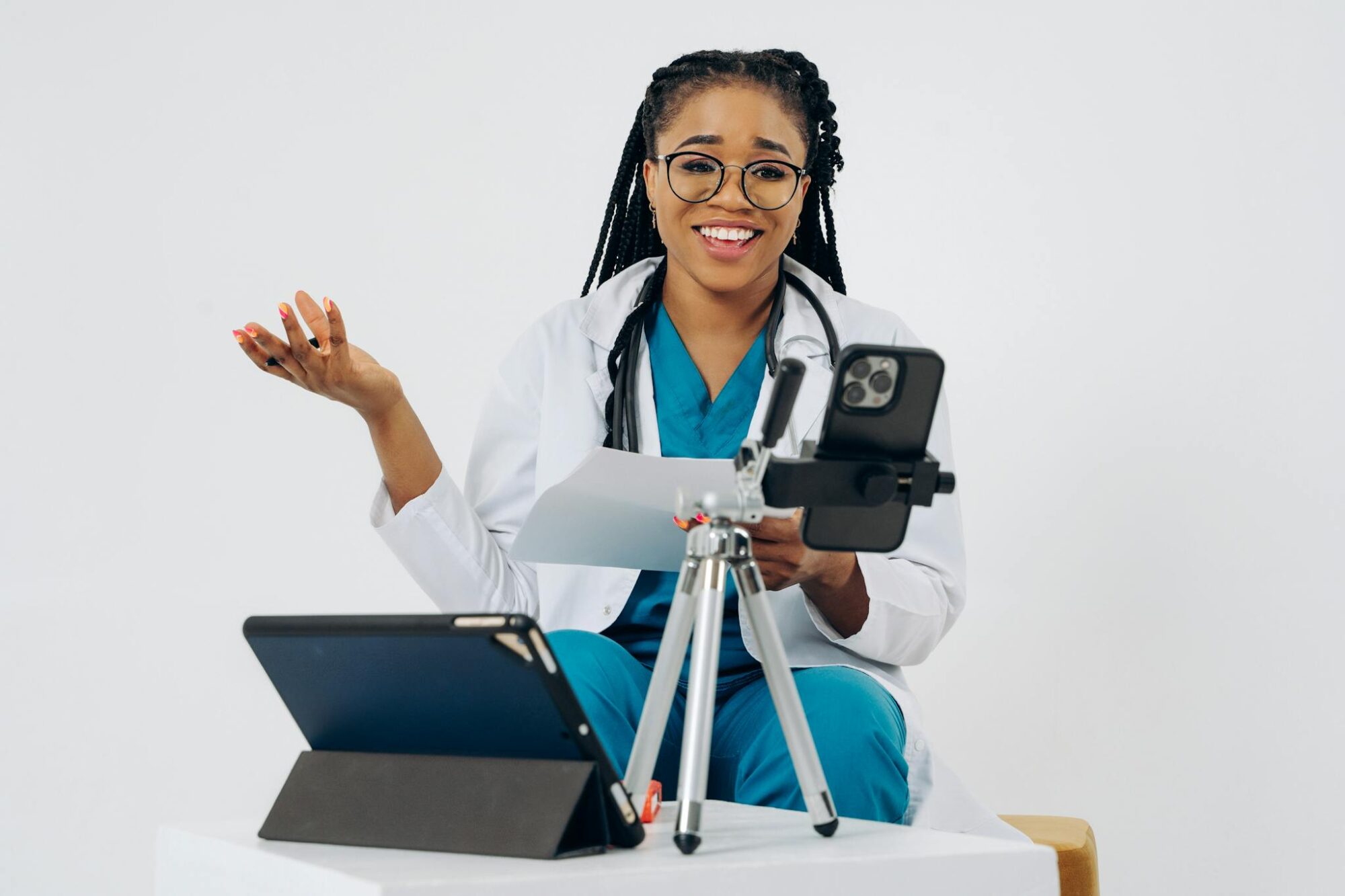 Black female doctor in virtual consultation, smiling at screen with stethoscope, tablet, and phone.