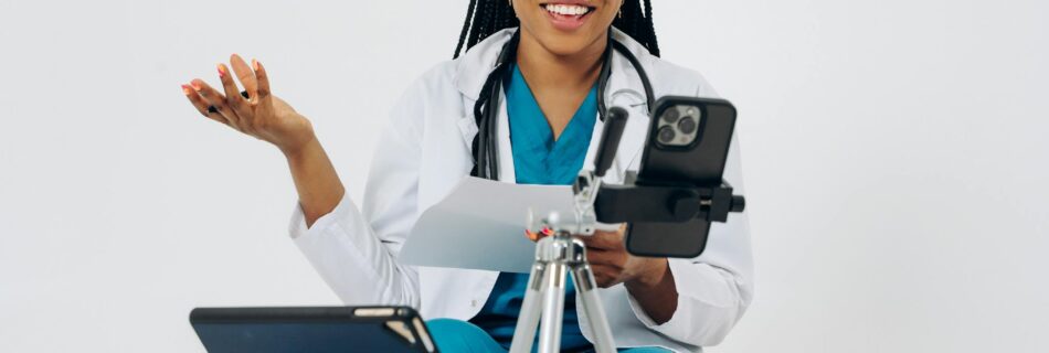 Black female doctor in virtual consultation, smiling at screen with stethoscope, tablet, and phone.